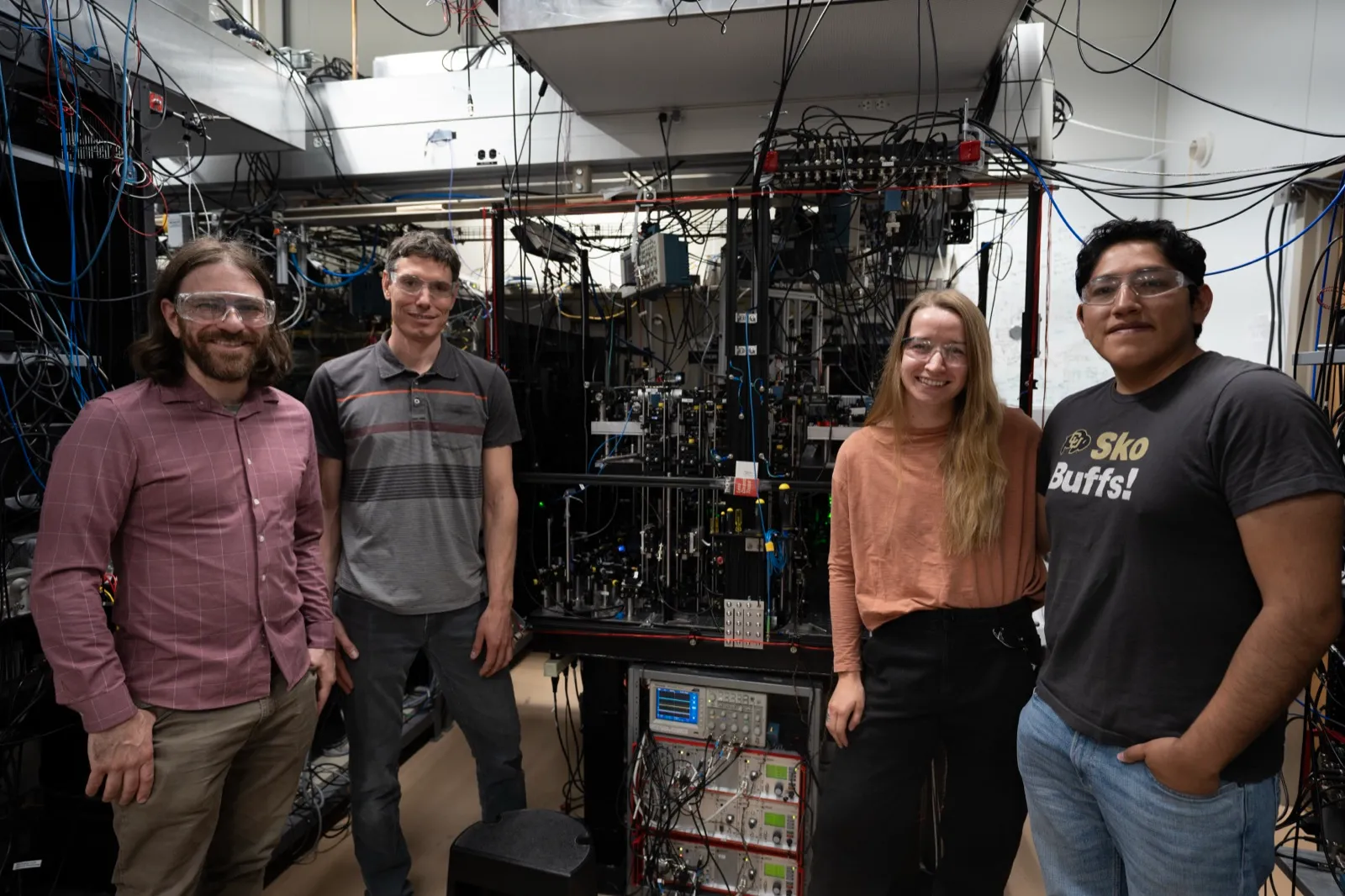 The NIST aluminum ion clock: Mason Marshall, David Hume, Willa Arthur-Dworschack, and Daniel Rodriguez Castillo. As of July 2025, this is the most accurate clock ever built. Image: R. Jacobson/NIST, public domain. The NIST aluminum ion clock team standing beside the apparatus — a complex array of lasers, optics, and electronics filling a large optical table